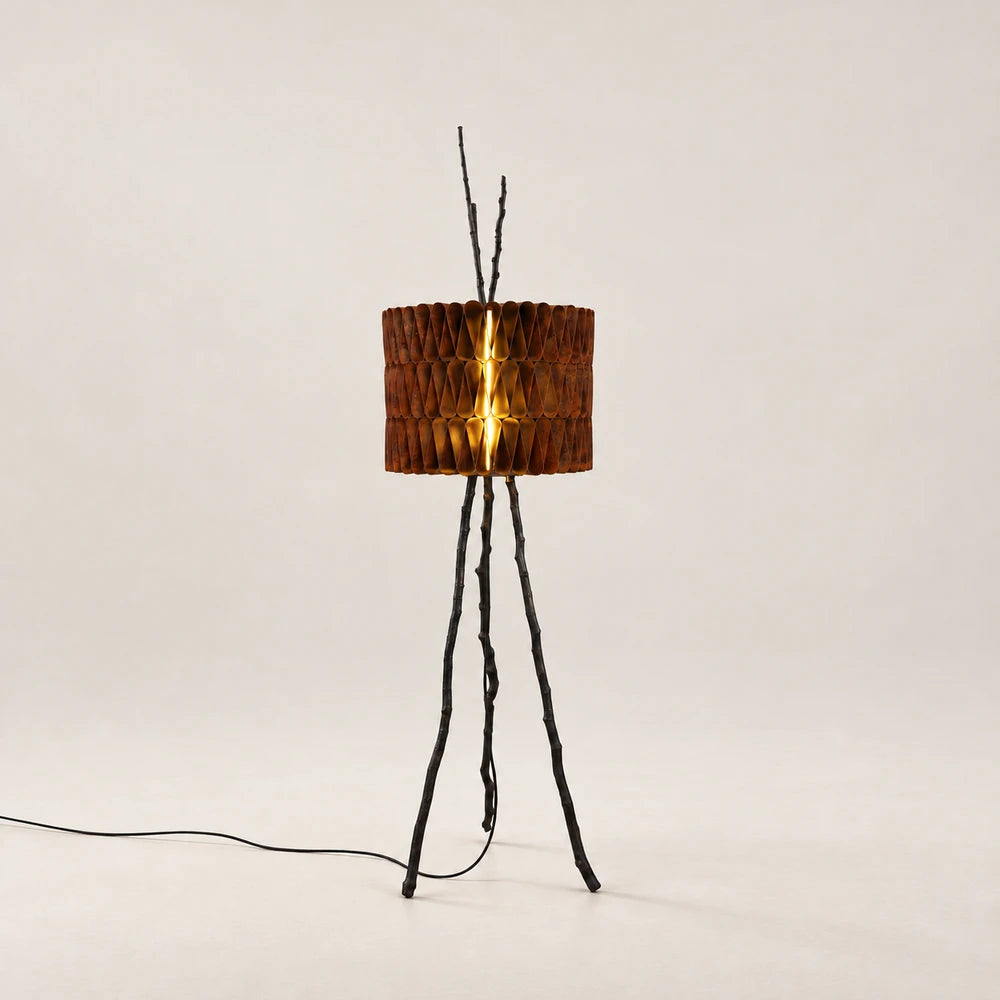 Tudor Floor Lamp by Eva Szumilas, with three dark branch-like legs and a cylindrical shade of folded rust-toned metal illuminated from within.